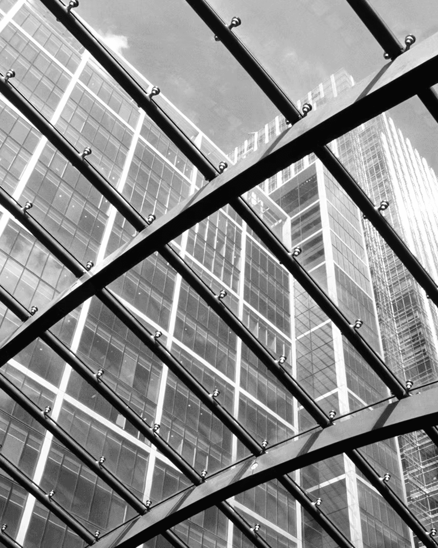 A high-contrast black-and-white architectural photograph capturing a geometric glass and steel canopy structure viewed from below, with diagonal metal beams intersecting to form a grid pattern that frames the towering glass-facade skyscrapers rising into a cloudy sky in the background, emphasizing modern urban design and structural symmetry.