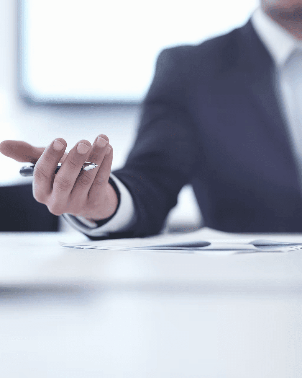 A close-up of a man's hand holding a sleek metallic pen, positioned over a white desk with scattered documents, while wearing a dark suit and white shirt, suggesting a professional legal or business setting with soft natural lighting from a blurred window in the background.
