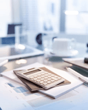 A close-up of a modern office desk featuring a sleek silver calculator resting on a stack of printed documents with visible bar charts and text, alongside a white pen and a brown notebook; in the softly blurred background, a white coffee cup on a saucer, a glass of water, and a laptop are subtly visible near a bright window, suggesting a professional, analytical, or financial workspace with a clean, minimalist aesthetic.