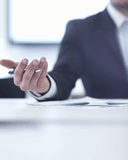 A close-up of a man's hand holding a sleek metallic pen, positioned over a white desk with scattered documents, while wearing a dark suit and white shirt, suggesting a professional legal or business setting with soft natural lighting from a blurred window in the background.